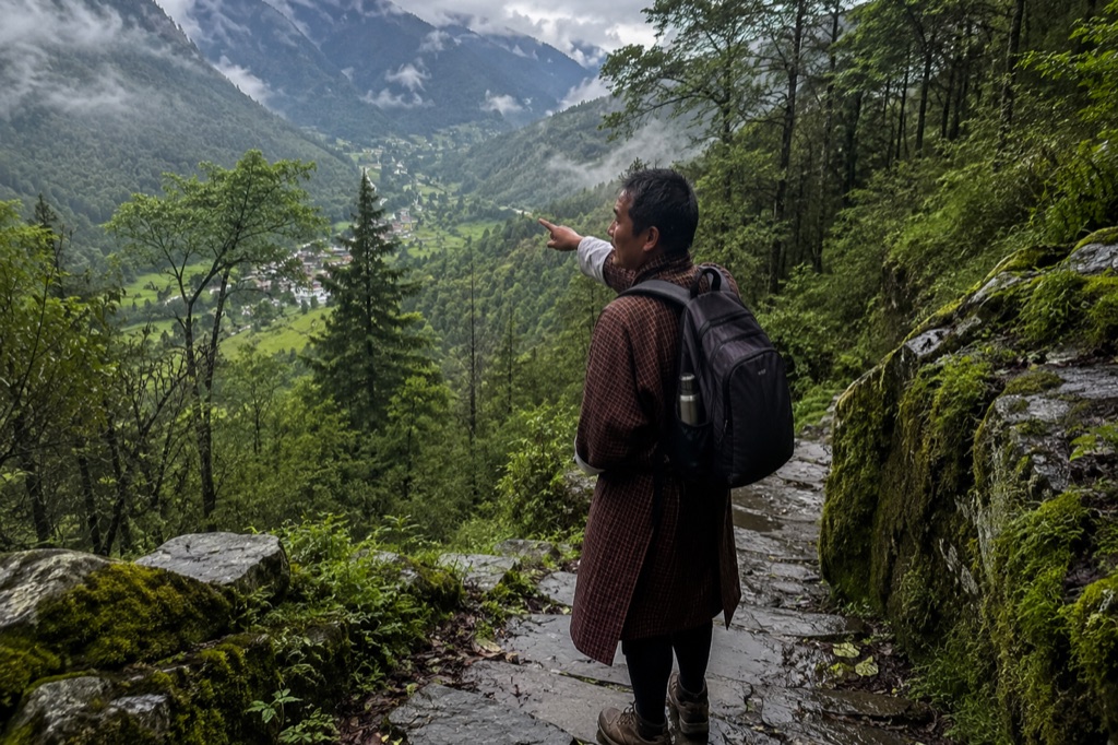 A guide stopped on a stone path on a forested slope, pointing up the valley.