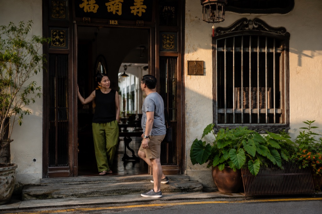 Two people talking in a shophouse doorway in late afternoon light.