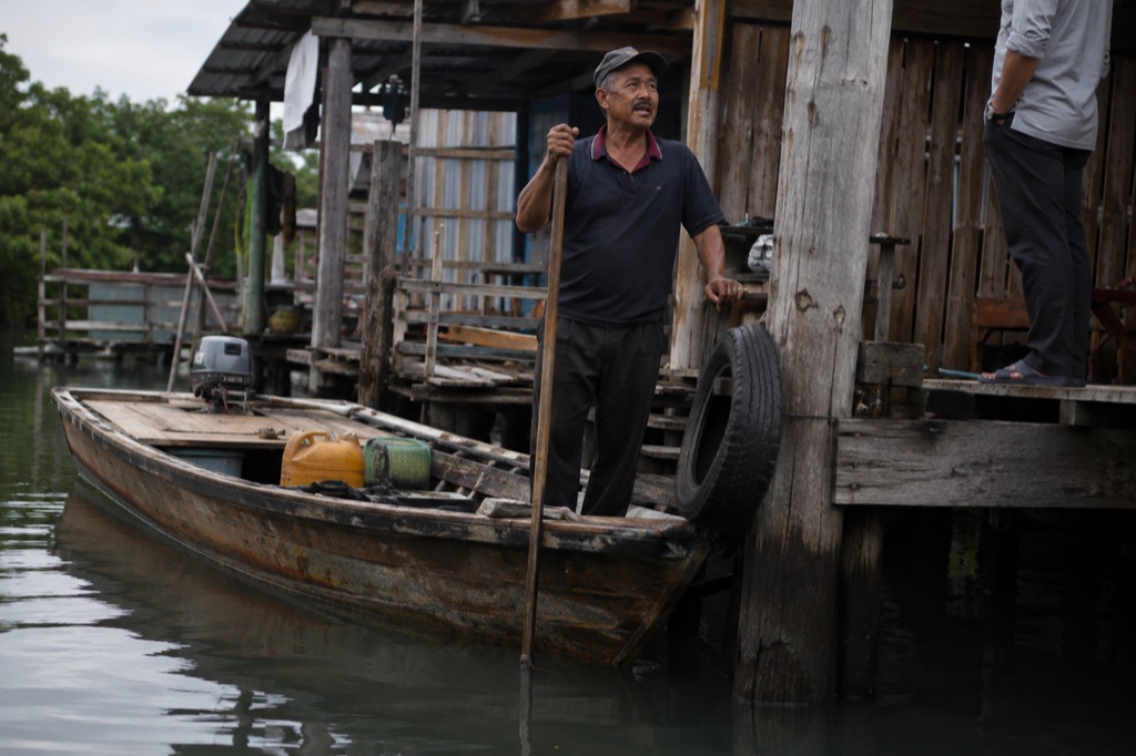 A boatman standing in a long wooden boat at a jetty, talking to somebody on the dock.