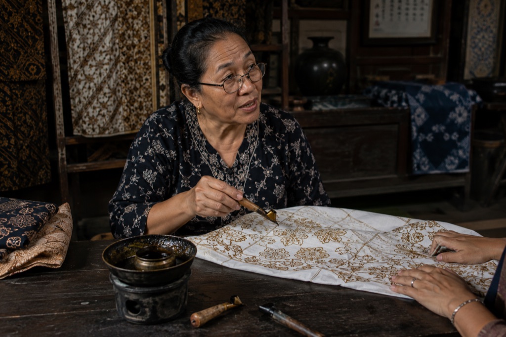 A woman applying wax with a canting at a batik table, explaining as she works.